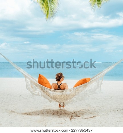 Young woman relaxing in wicker hammock on the sandy beach on Mauritius coast and enjoying wide ocean view waves. Exotic countries vacation and mental health concept image. Royalty-Free Stock Photo #2297861437