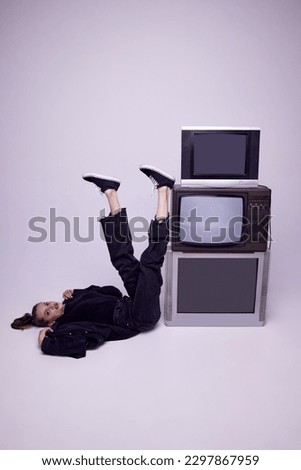 Beautiful young girl in jeans clothes lying on floor near retro tv sets over grey studio background. Television propaganda. Concept of fashion, 80s, 90s style, retro and vintage, mass media
