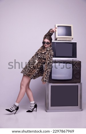 Creative translation. Young girl, influencer in stylish coat and trendy sunglasses posing near retro TV sets against grey background. Concept of fashion, 80s, 90s style, retro and vintage, mass media