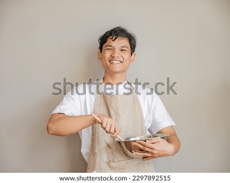 Beautiful many wearing casual white t-shirt standing in front of grey background showing half body shot of young southeast asian burmese man, cooking apron kitchen chef baker wooden spoon
 Royalty-Free Stock Photo #2297892515