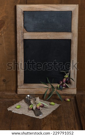 Black chalkboard for menu, spoon and fork at napkin and fresh olives over wooden background.