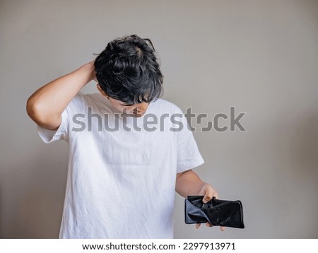 Beautiful many wearing casual white t-shirt standing in front of grey background showing half body shot of young southeast asian burmese man, checking wallet for money issue broke empty wallet Royalty-Free Stock Photo #2297913971