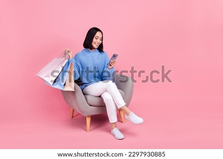 Full length photo of cute girl blue sweater white pants look at smartphone hold clothes sit on chair isolated on pink color background