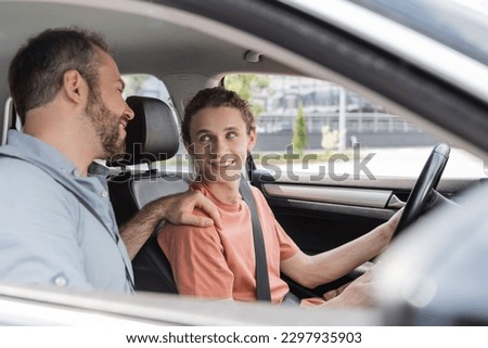cheerful father putting hand on shoulder of teenage son while teaching him how to drive car Royalty-Free Stock Photo #2297935903