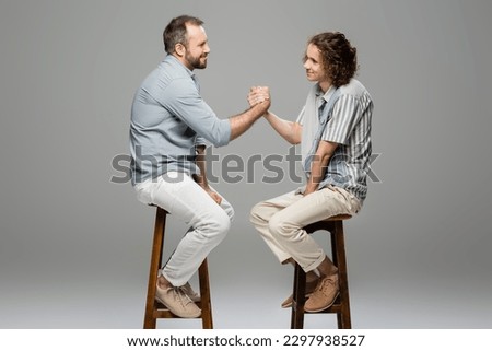 side view of happy father and teenage son shaking hands while sitting on high chairs on grey Royalty-Free Stock Photo #2297938527