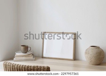Empty horizontal picture frame mockup in minimalist interior. Wooden table, blurred rattan chair. Vase with cup of coffee. White wall background. Landscape template of artwork, painting, poster.