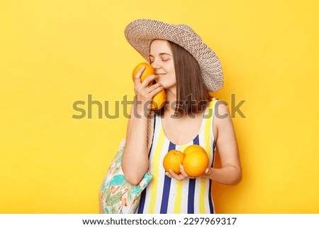 Enjoyable calm woman wearing striped swimsuit and straw hat isolated yellow background standing holding fresh fruit smelling aromat of oranges.