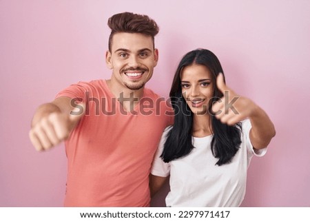 Young hispanic couple standing over pink background approving doing positive gesture with hand, thumbs up smiling and happy for success. winner gesture. 