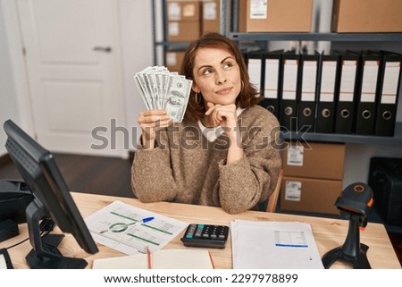 Young beautiful woman working at small business ecommerce holding money serious face thinking about question with hand on chin, thoughtful about confusing idea 