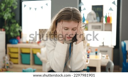 Young blonde woman preschool teacher stressed sitting on chair at kindergarten