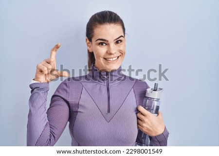 Beautiful woman wearing sportswear holding water bottle smiling and confident gesturing with hand doing small size sign with fingers looking and the camera. measure concept.  Royalty-Free Stock Photo #2298001549