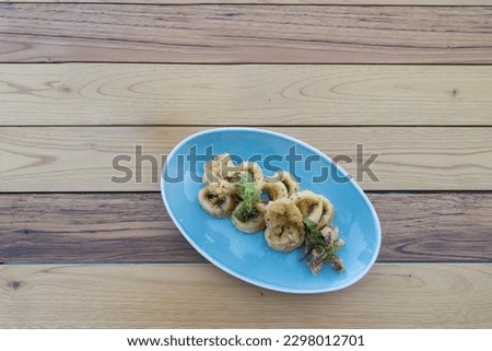 Isolated blue plate of breaded calamari rings, typically known by "calamares a la andaluza" in Spain, with edible green seaweeds on top. Picture of Mediterranean food on wooden background.