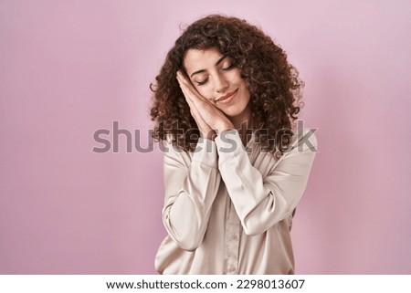 Hispanic woman with curly hair standing over pink background sleeping tired dreaming and posing with hands together while smiling with closed eyes. 