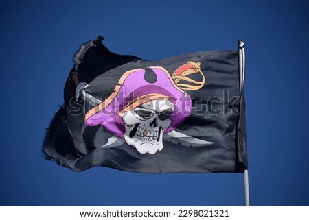 Black pirate flag with a pirate skull in a purple hat and swords flapping in the wind against a clear, dark blue sky.