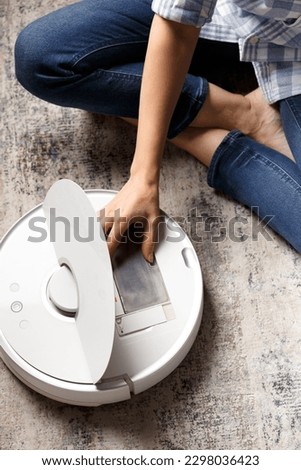 The girl takes the dust container out of the vacuum cleaner robot to clean it. The concept of a smart home, housework, cleaning, keeping the apartment clean, smart technologies. Royalty-Free Stock Photo #2298036423