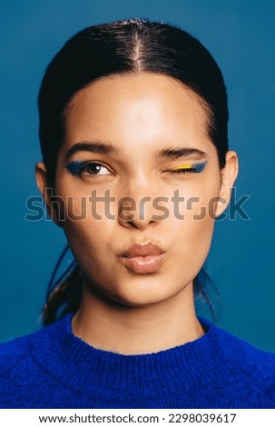 Young woman with bold eyeliner winks and pouts her lips, making a flirty expression. Radiant young woman wearing vibrant makeup and a blue sweater in a studio. Royalty-Free Stock Photo #2298039617