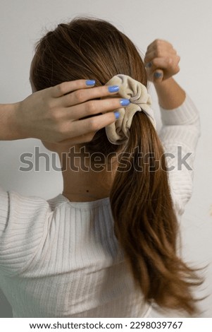 Young, blonde woman touching beige scrunchie on her sleek ponytail. Hands in movement, arms raised. Model against white background. Royalty-Free Stock Photo #2298039767