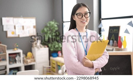 Young chinese woman preschool teacher smiling confident holding clipboard at kindergarten