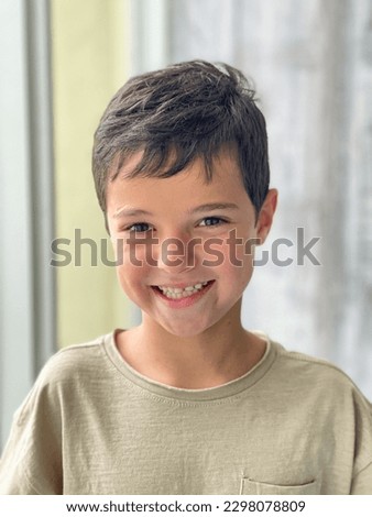 Portrait of a cheerful positive child with a dental smile. Caucasian brunette boy smiling while standing at the window. The concept of happiness, childhood, joy. Eye contact. Indoor. Royalty-Free Stock Photo #2298078809