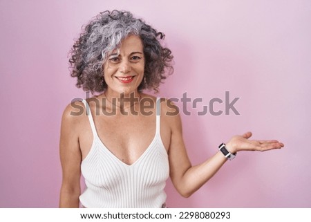 Middle age woman with grey hair standing over pink background smiling cheerful presenting and pointing with palm of hand looking at the camera. 