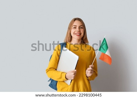Young woman with flag of Ireland, laptop and backpack on grey background