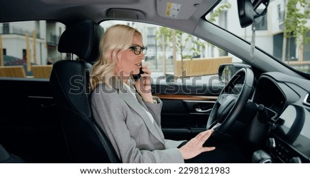 Attractive stylish woman sitting in car dressed in formal clothes and glasses talking on cell phone , street background off car window