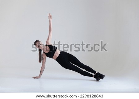 A young girl with brown hair, with a tattoo in a black top and leggings on a white background. Sports, fitness, is in the sidebar. Press exercises. Yoga Royalty-Free Stock Photo #2298128843