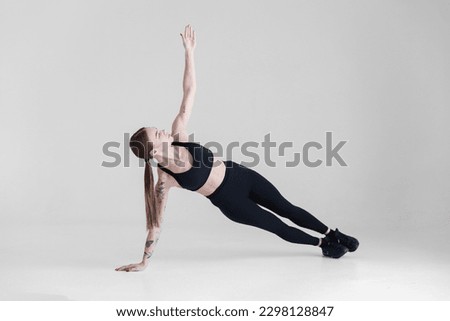 A young girl with brown hair, with a tattoo in a black top and leggings on a white background. Sports, fitness, is in the sidebar. Press exercises. Yoga Royalty-Free Stock Photo #2298128847
