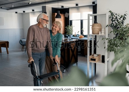 Lovely senior couple in retirement at home. Supportive wife taking care of husband with physical disability, helping him to walk around with mobility walker. Happy elderly couple in love.