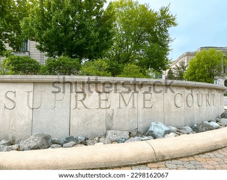 Engraved Stone in Front of the Illinois Supreme Court House that reads, "Illinois Supreme Court". Courthouse building stands behind the signage and green trees. Located in Springfield, IL, USA.