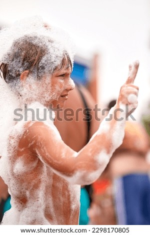 Happy little boy with foam on his head having fun in a water park, in wet swimwear at a foam party or holiday on a sunny hot summer day