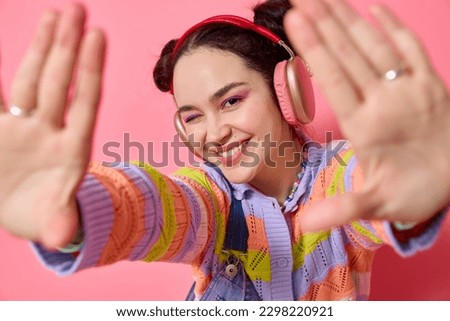 Close up young girl with buns in headphones listens to music  wears coloured knitted sweater looking at camera smiling and winking pov holding camera with hands isolated on seamless pink background. 