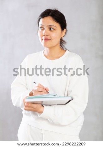 Portrait of smiling young Asian woman, tutor or college student, writing in workbooks while standing on gray studio background