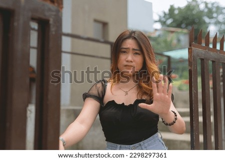 A lovely young lady standing by the gate looking serious and anxious with a furrowed forehead while gesturing wait. Restraining order at her house. Royalty-Free Stock Photo #2298297361