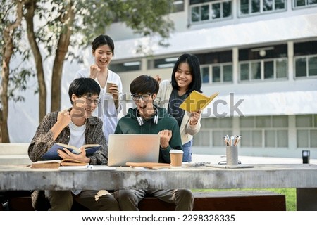 Group off cheerful young Asian college students looking at laptop screen, rejoicing, and celebrating their school project success or exam score together at a campus park.