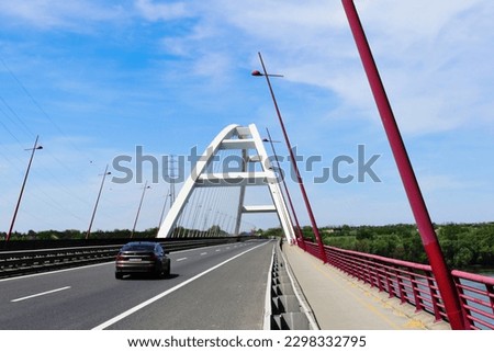 white basket-handle arch design steel cable suspension bridge. blue sky. high voltage electrical cables with red cable marker balls for aviation safety. diminishing perspective. red street lights.