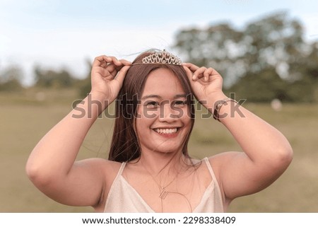A beautiful young adult woman wearing a bright smile on her face as she celebrates her 20th birthday. She has adorned a tiara on her head, signifying her special day. Green nature background.