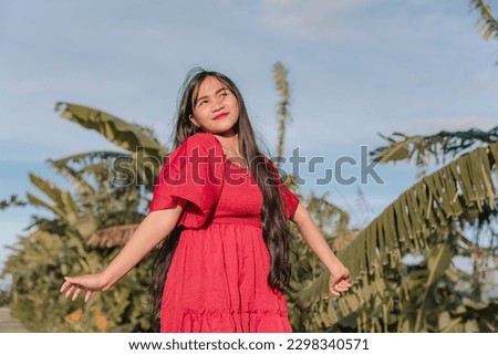 A charming lady wearing a long red dress , smiling sweetly, gazes at the sky while posing being free and confident of herself. Clear sky and trees in the background