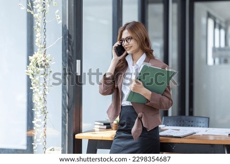 Asian Woman entrepreneur busy with documents her work in the office. Young Asian woman talking over smartphone or cellphone while working on computer at her desk.