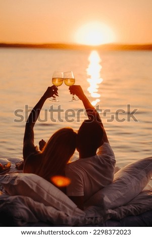 Silhouettes of a happy couple raising glasses on a summer evening near the sea at sunset. Royalty-Free Stock Photo #2298372023