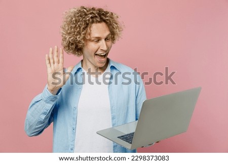 Young fun blond IT man wears blue shirt white t-shirt hold use work on laptop pc computer hold use work on laptop pc computer waving hand speak get video call isolated on plain pastel pink background