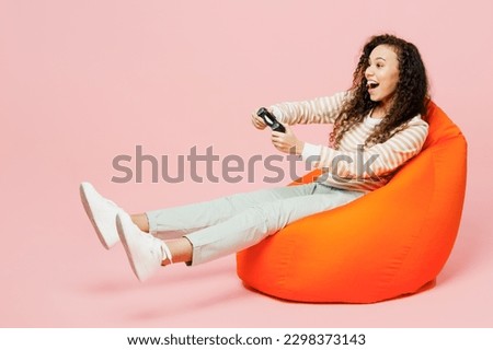 Full body gambling young woman of African American ethnicity she wear light casual clothes sit in bag chair play pc game with joystick console isolated on plain pastel pink background studio portrait