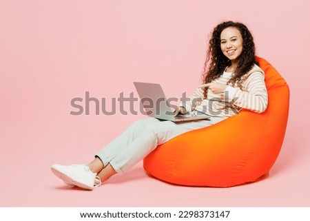 Full body young IT woman of African American ethnicity she wear light casual clothes sit in bag chair hold use work poitn finger on laptop pc computer isolated on plain pastel pink background studio