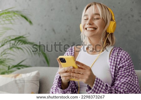 Satisfied smiling young woman wearing casual clothes headphones listen music use mobile cell phone sits on grey sofa couch stay home hotel flat rest relax spend free spare time in living room indoor Royalty-Free Stock Photo #2298373267