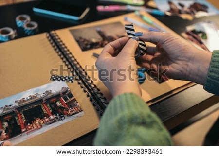 Middle-aged woman's hands holding a roll of washi tape that she is going to use to paste a photo onto her handmade kraft travel album. The sign says the word temple in Thai languaje. Royalty-Free Stock Photo #2298393461