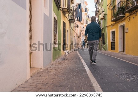 Back picture of gray-haired gentleman walking his white curly haired dog through colorful streets.