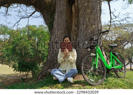 Women sitting under tree and covering half face with a book while resting after cycling in the park.