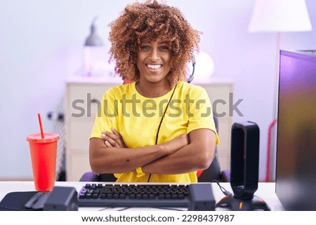 Young hispanic woman with curly hair playing video games wearing headphones happy face smiling with crossed arms looking at the camera. positive person. 