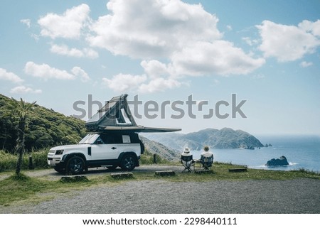 Couple sitting in a camping chair in nature with a view of Island in a beautiful blue sky with an overlanding car with a rooftop tent and an awning.