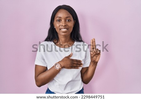 African young woman wearing casual white t shirt smiling swearing with hand on chest and fingers up, making a loyalty promise oath  Royalty-Free Stock Photo #2298449751